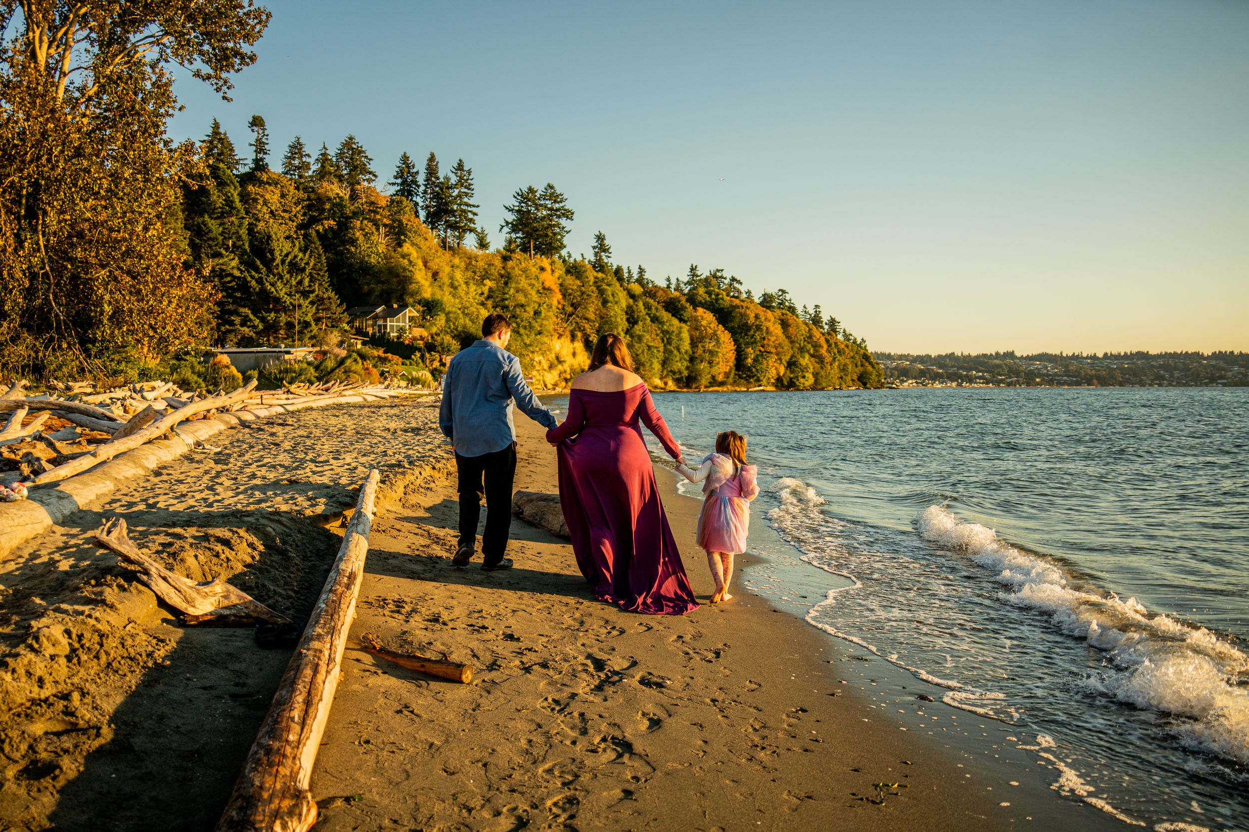 A family walks together along a beach at sunset with evergreen trees lining the shore.