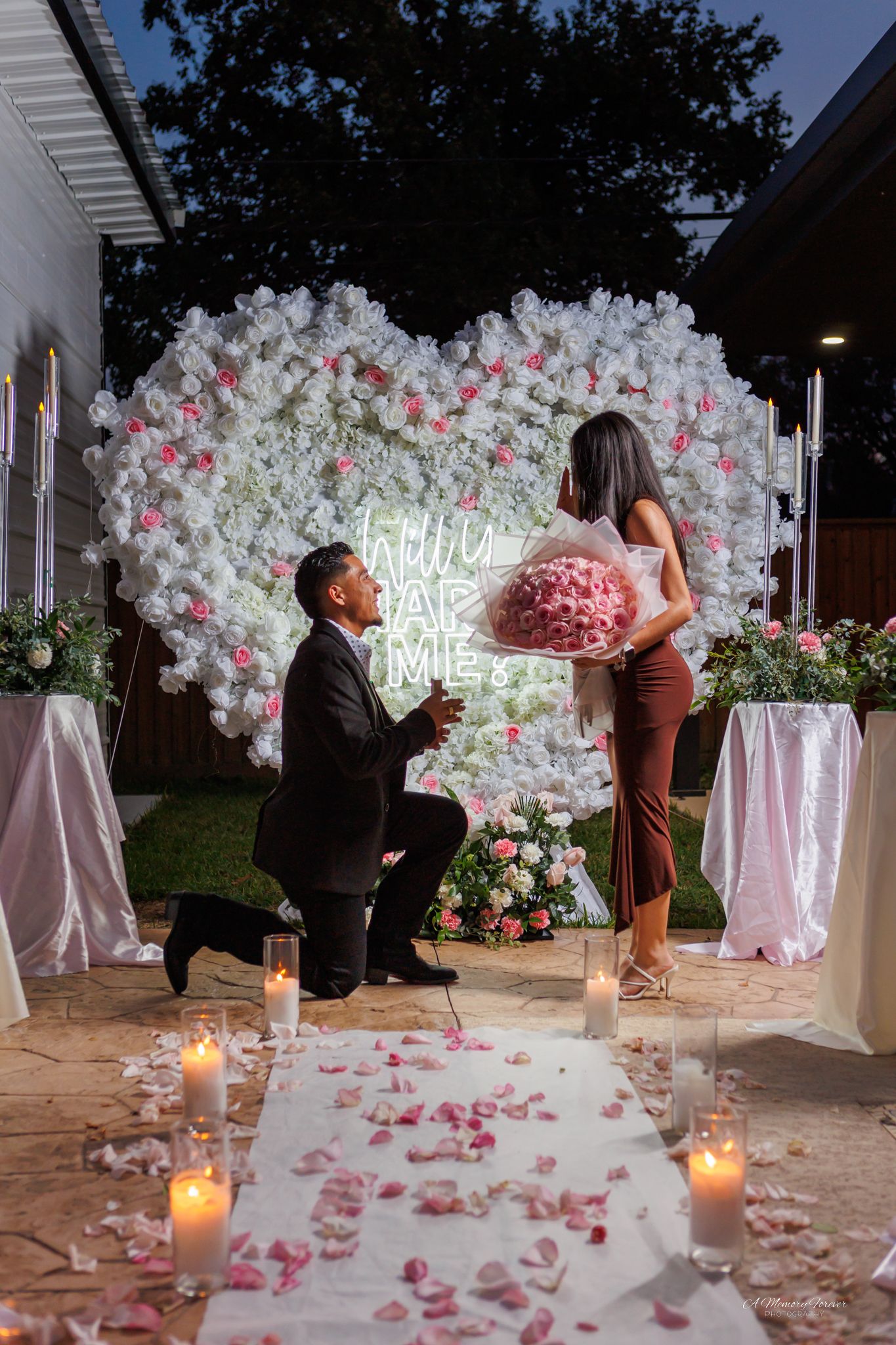 Man proposes on one knee before a floral heart backdrop with Will You Marry Me neon sign, candles, and rose petals.
