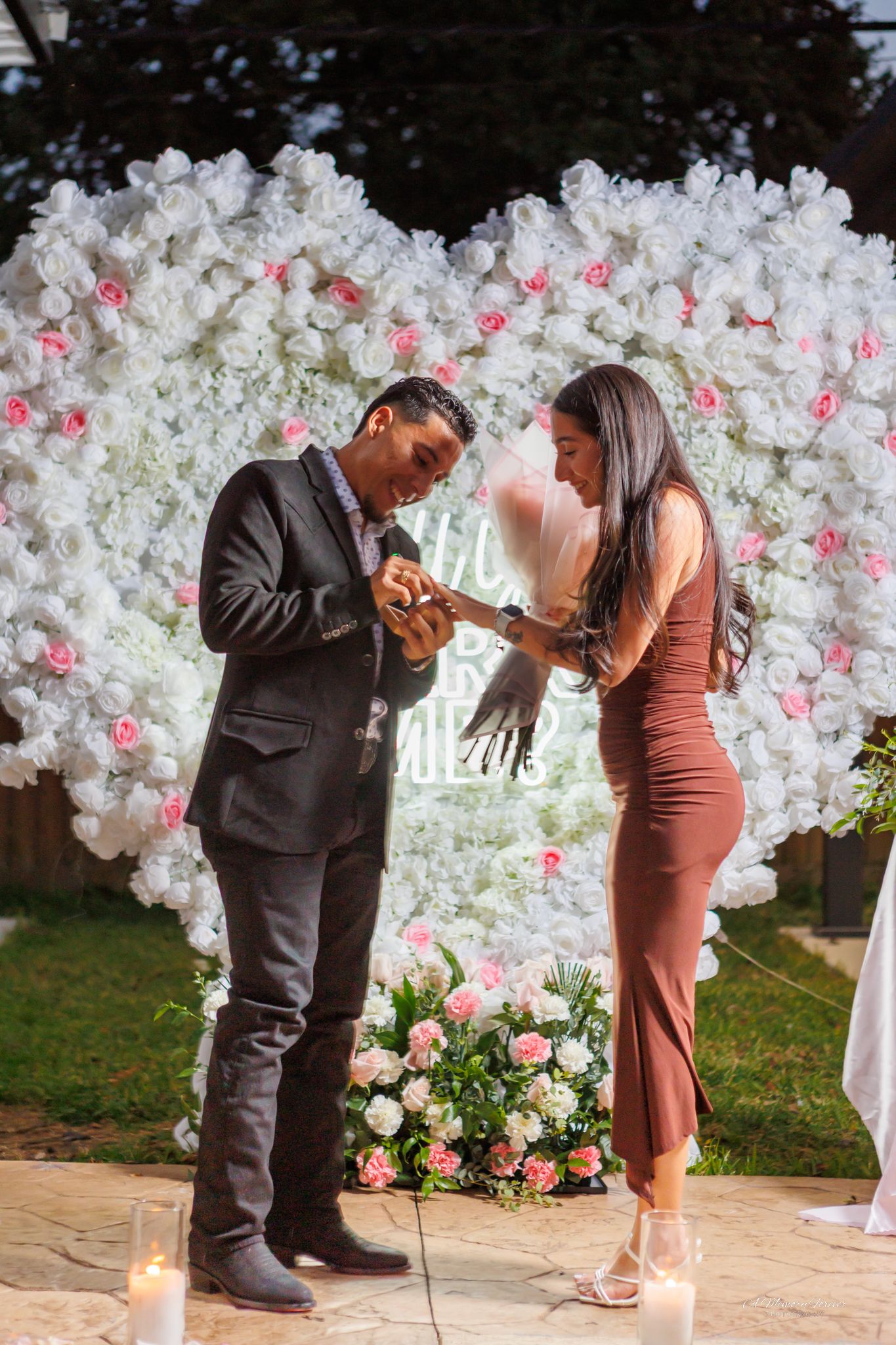 Man placing engagement ring on woman's finger in front of white floral heart backdrop with pink roses and candles.