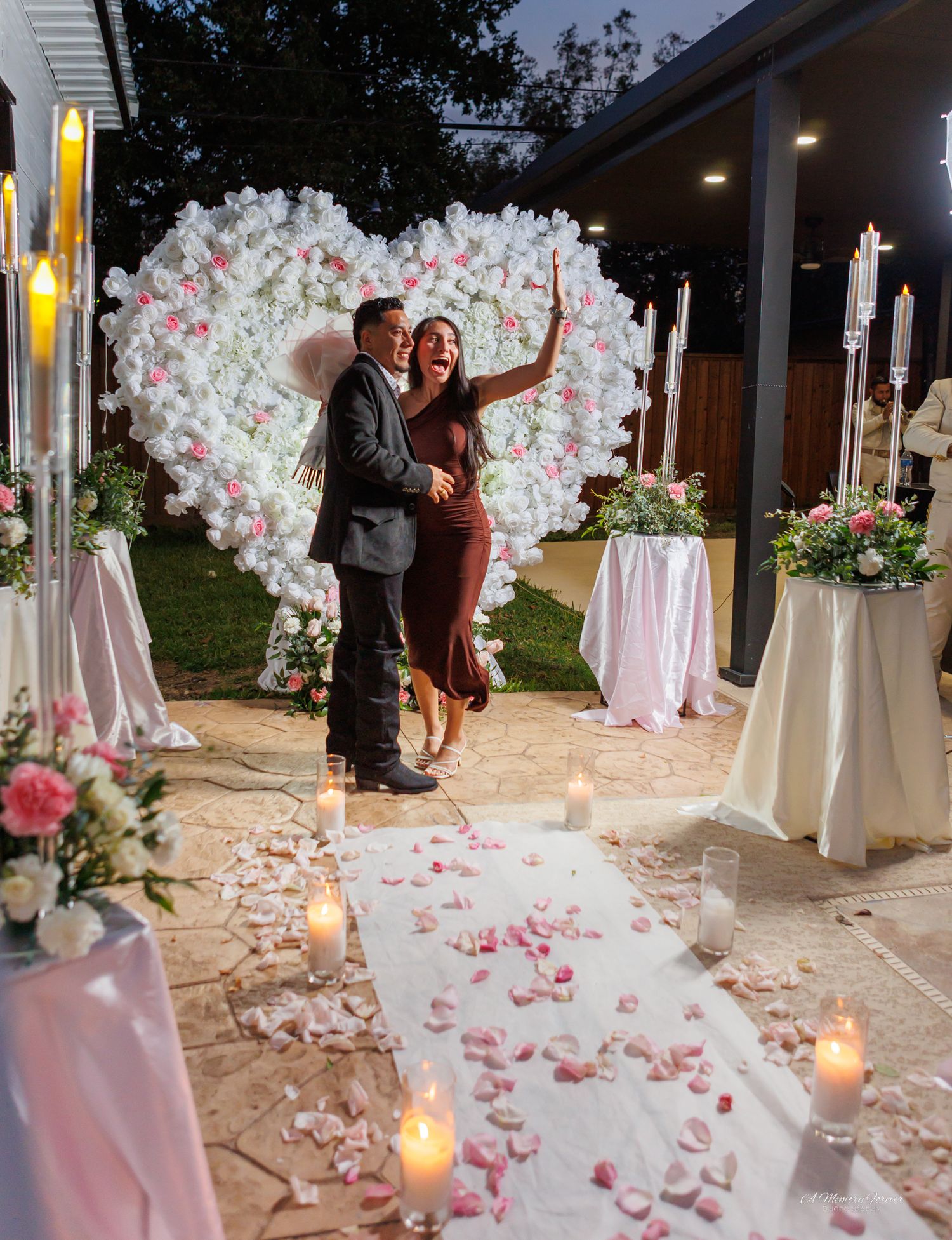 Couple celebrating a romantic proposal with a floral heart backdrop, rose petals, and candles at an outdoor evening event.