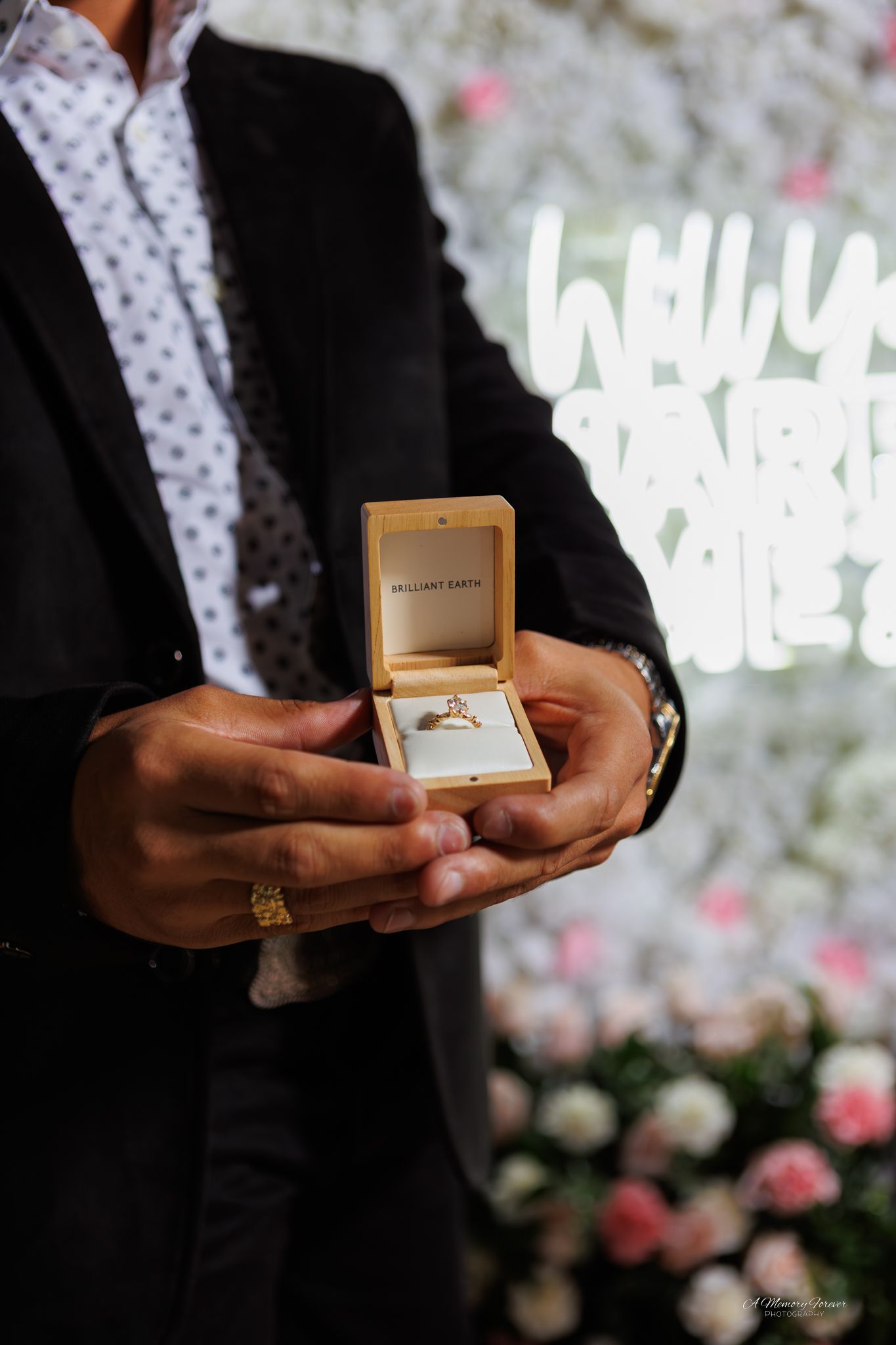 Man in black suit holding open Brilliant Earth ring box with gold diamond engagement ring, floral backdrop with neon sign.