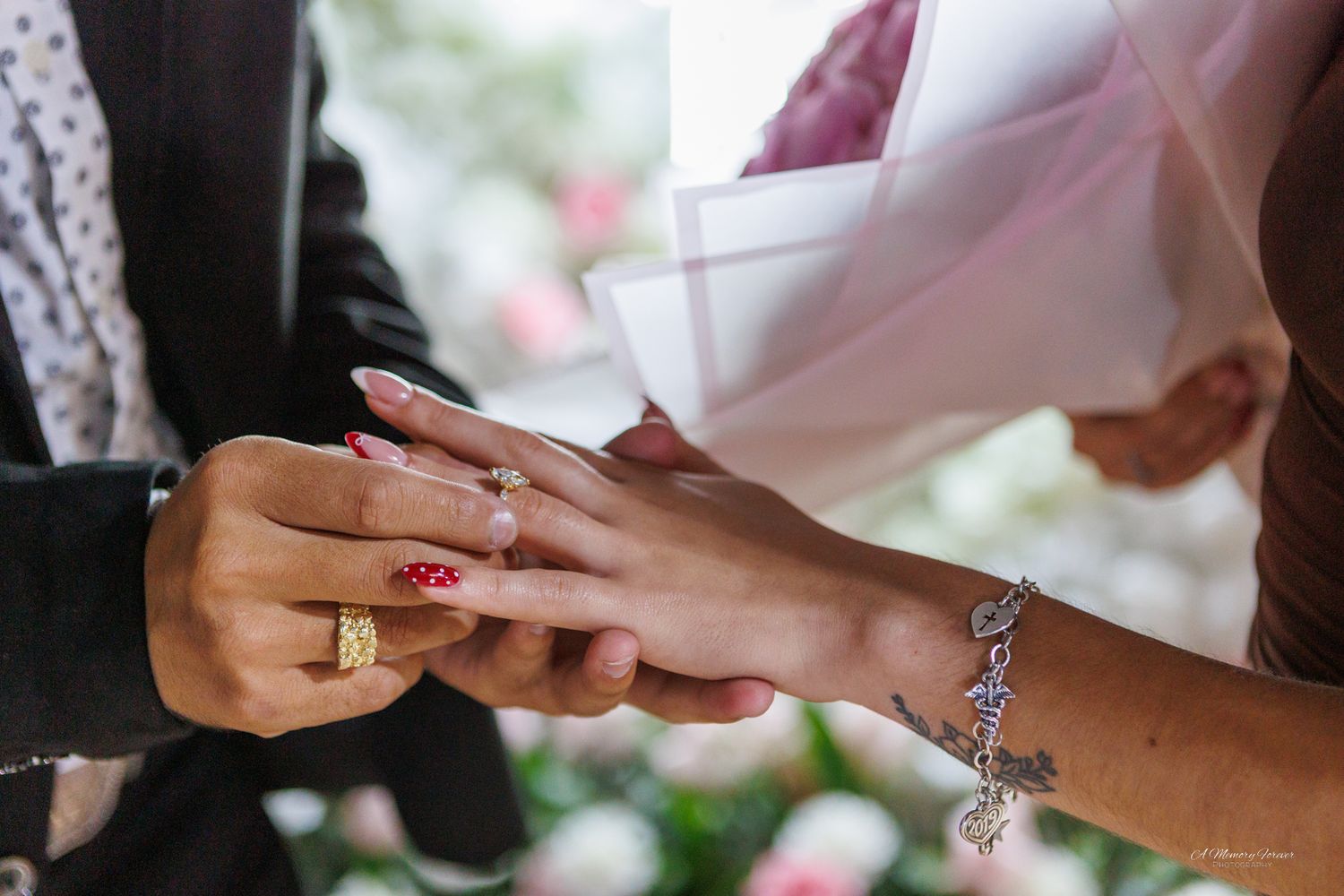 Groom placing a diamond wedding ring on bride's finger during ceremony, bride wearing charm bracelet and red nail polish.