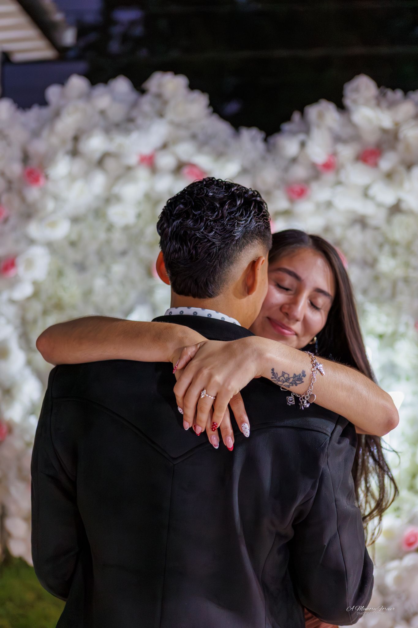 Couple embracing at wedding with white and pink floral backdrop, woman smiling with eyes closed showing engagement ring.