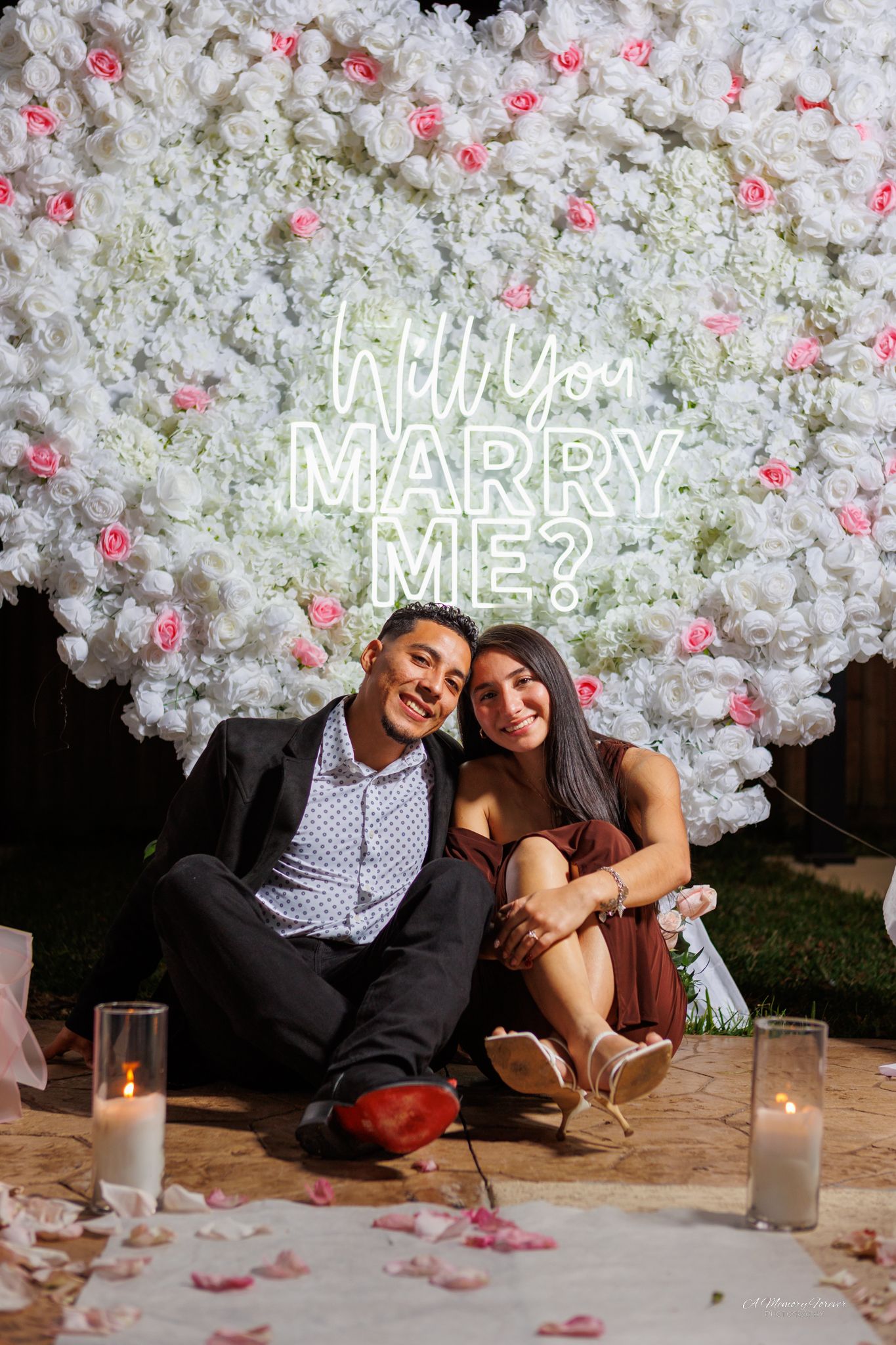 Happy couple sits together in front of a Will You Marry Me neon sign on a white floral wall after proposal.