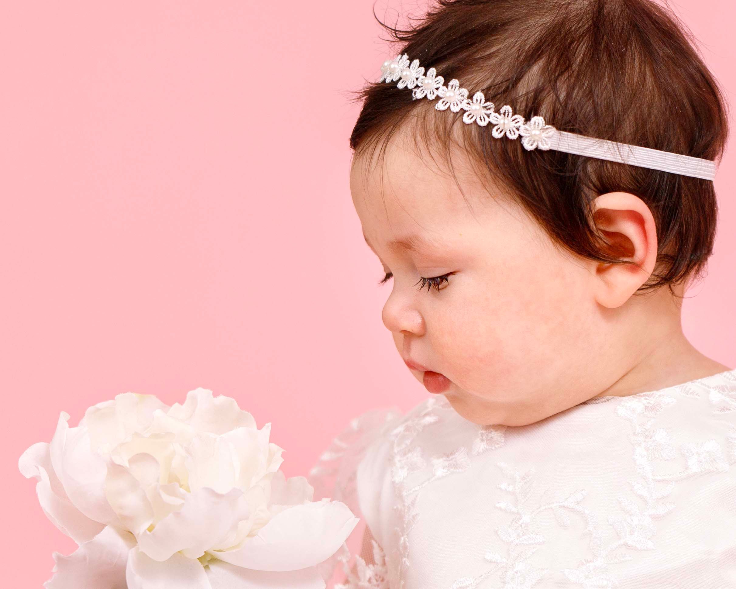 Bergen County Family Photographer - Christening photo of a baby looking at white flower.  In studio in front of pink background in Mahwah