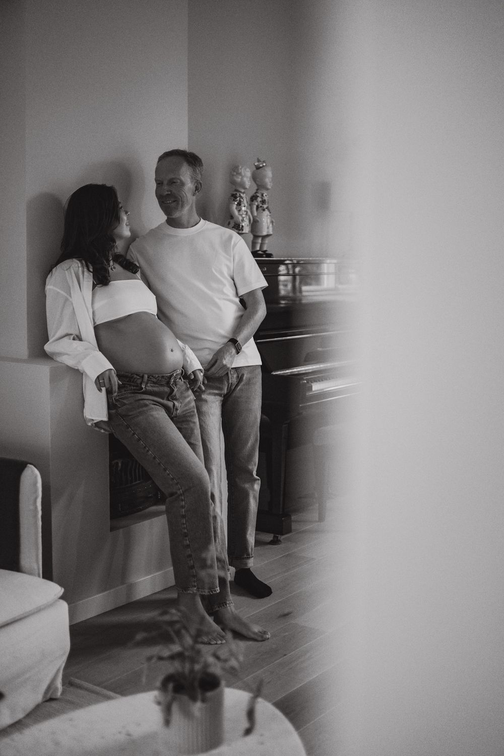 Black and white photo of a pregnant couple leaning against a wall, sharing a tender moment at home.