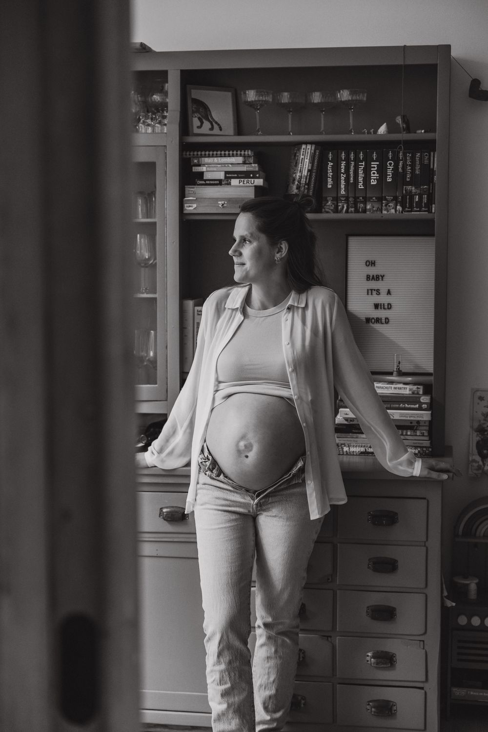 Black and white portrait of an expectant mother standing beside a bookshelf, wearing casual attire and showing her baby bump.