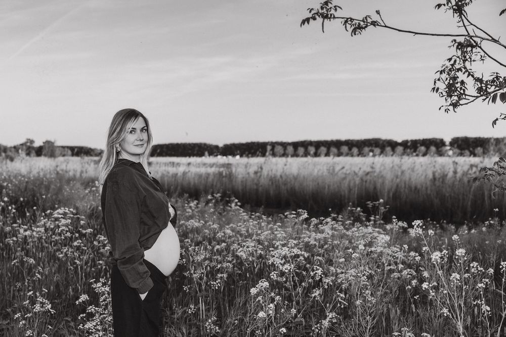 A pregnant woman stands in a black and white wildflower meadow at sunset, silhouetted against a peaceful countryside landscape.