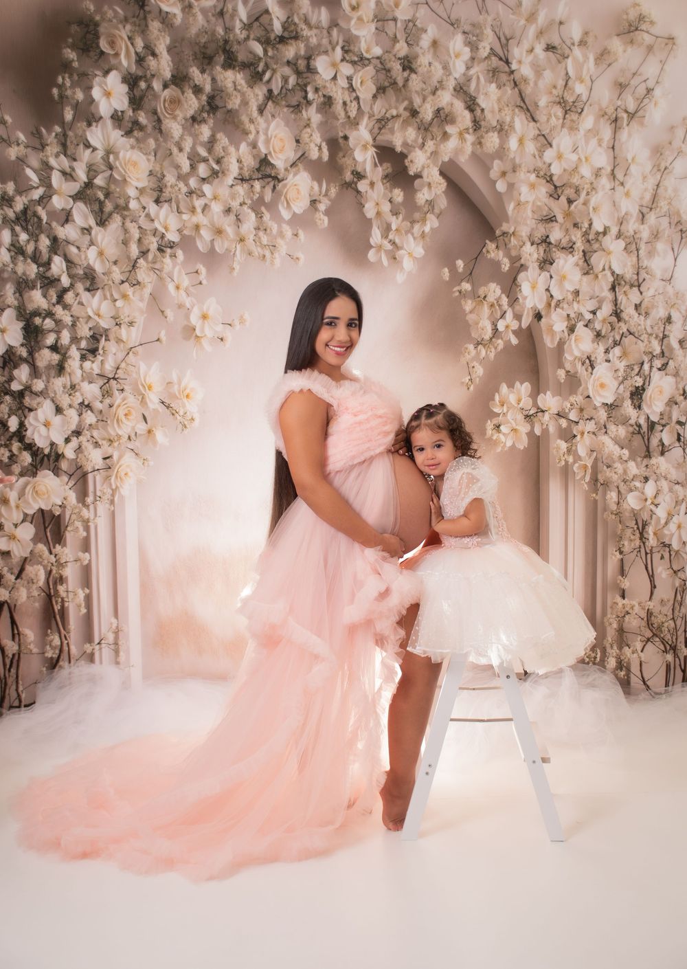 Ethereal maternity photo series in pink dress against white floral backdrop creating a dreamy romantic atmosphere.