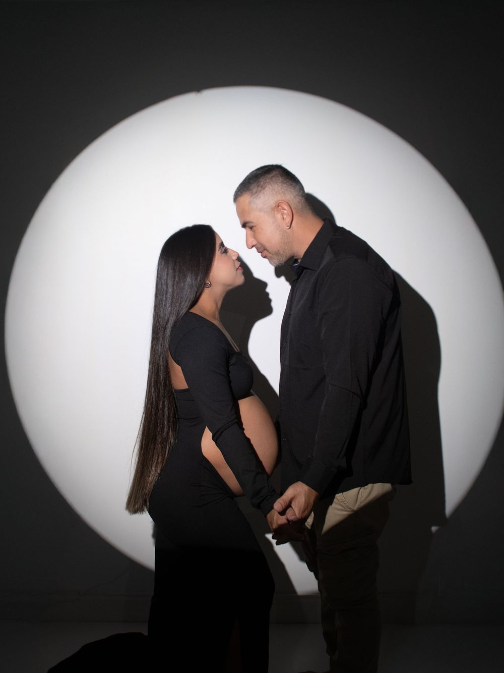 A romantic couple in black attire pose against a dramatic white circular backdrop in a dark studio setting.