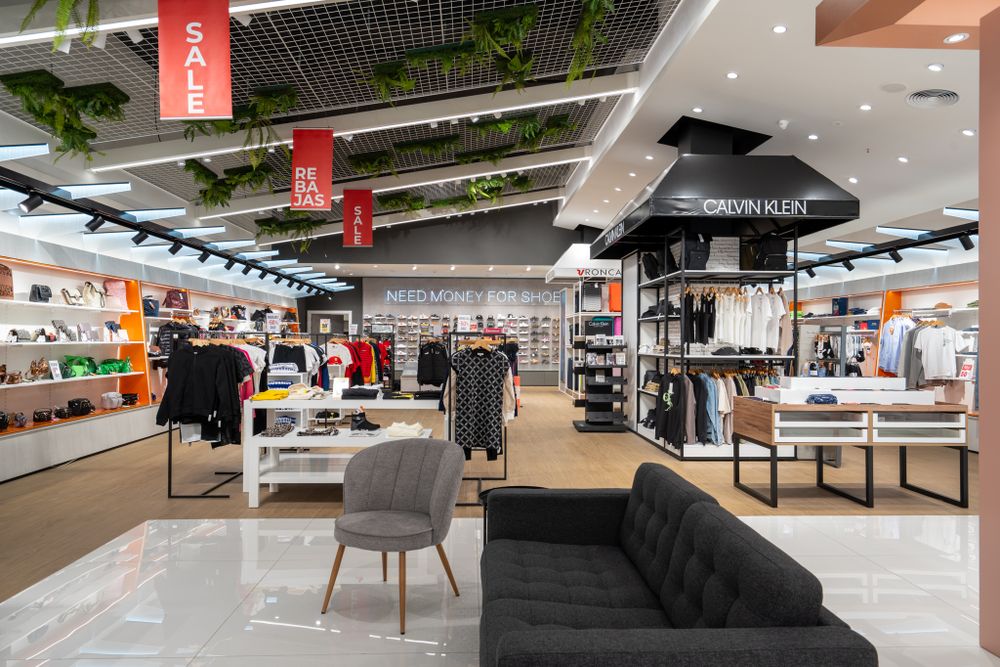 Spacious retail store interior with clothing displays, gray seating, and decorative ceiling elements.