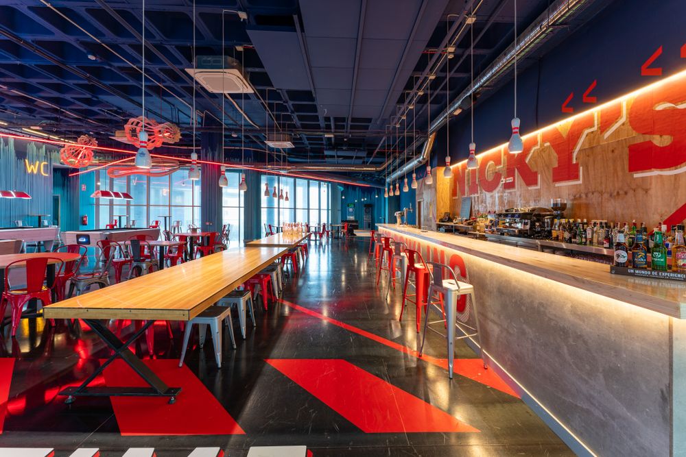 Modern restaurant interior with long communal table and bar featuring vibrant red lighting and industrial design elements.