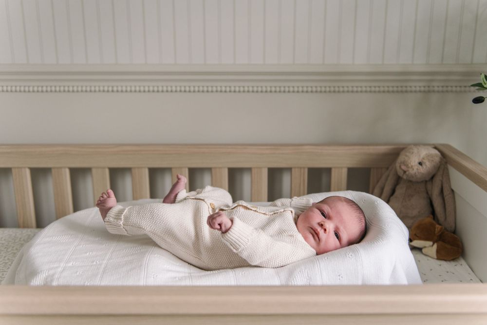A sleeping baby in white clothing lies on a mattress in a wooden crib with a plush bunny toy nearby.