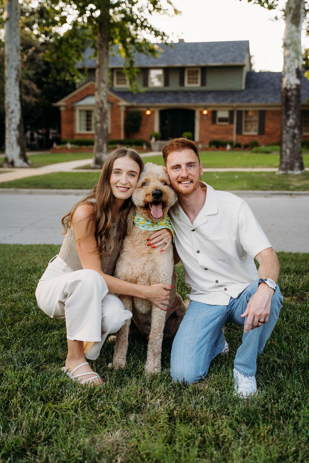 A goldendoodle dog poses with its owners on their front lawn in front of a brick house.