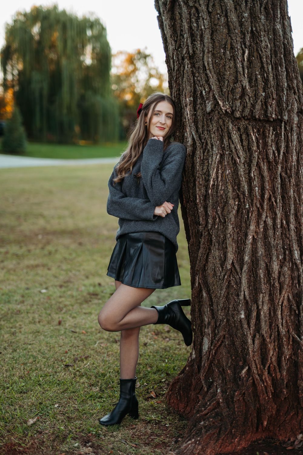 Person in dark sweater and skirt posing against large tree trunk in autumn setting.
