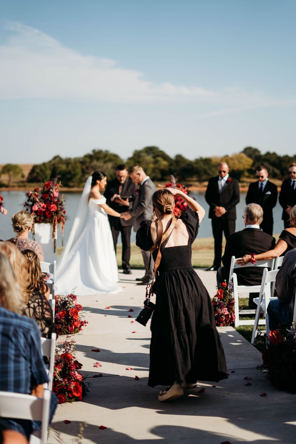 Outdoor lakeside wedding ceremony with red floral arrangements and white chairs.