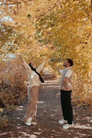 Couple in casual wear playfully reaching up towards golden fall leaves on a forest path.