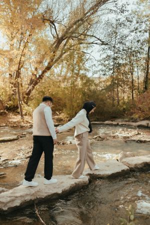 Couple holding hands while walking carefully across rocky creek bed in autumn setting.