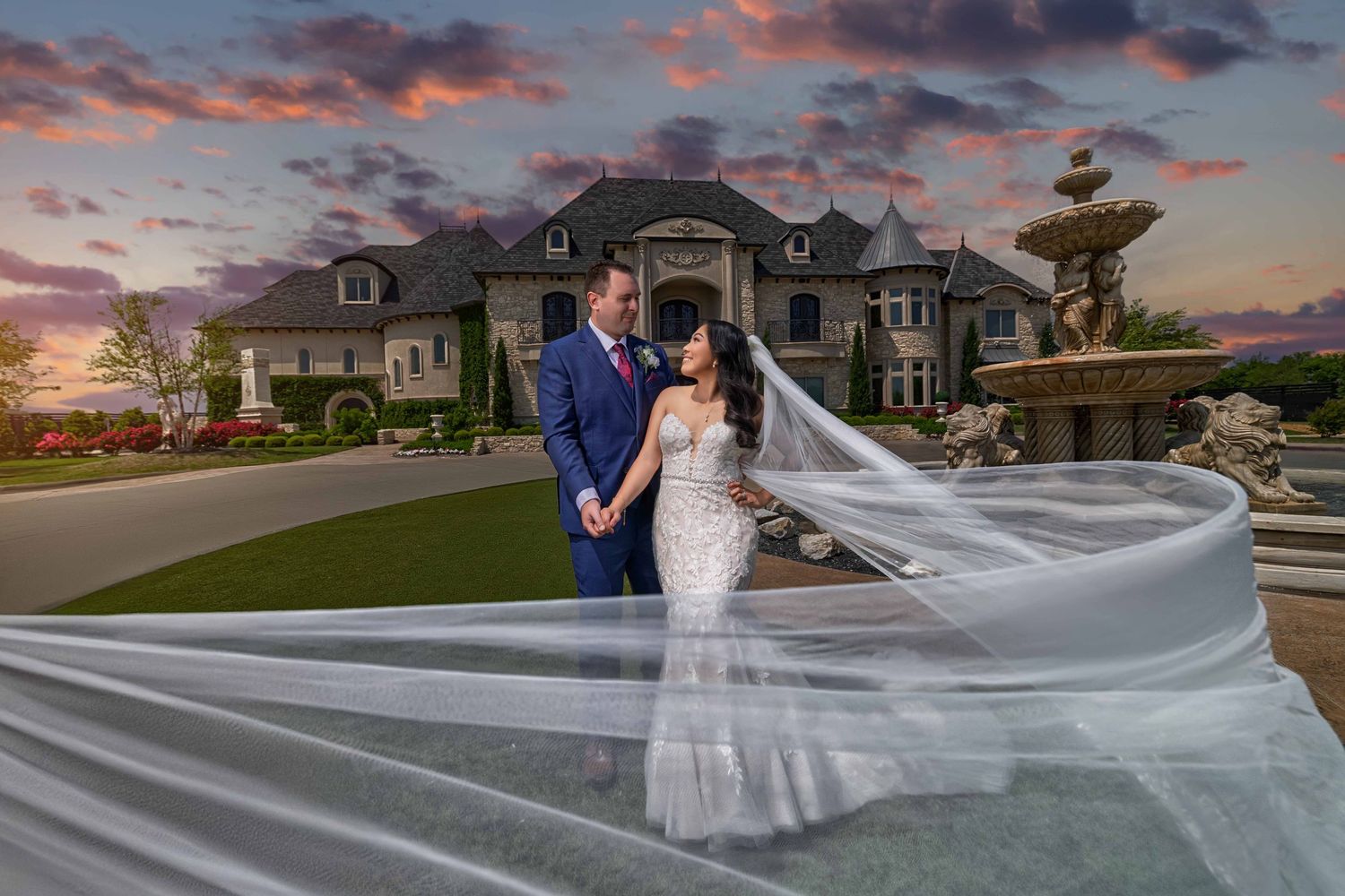 Bride and groom holding hands in front of a luxury stone manor at sunset with a long white veil caught in the wind by a wedding photographer