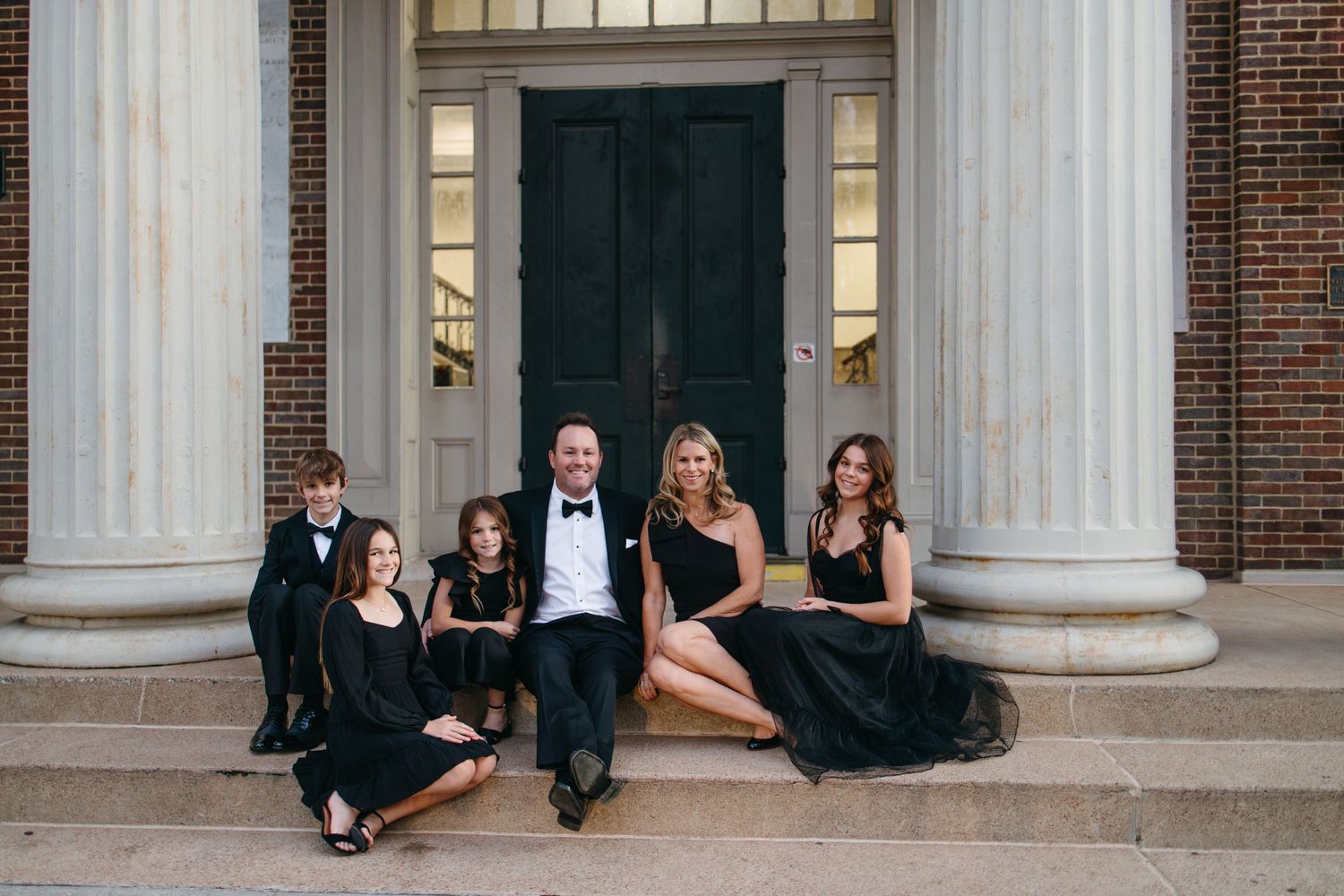 A formal group sits together on stone steps between grand white columns of a historic brick building.
