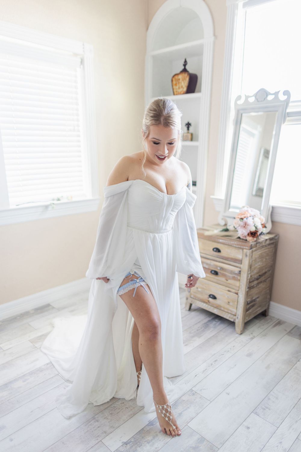 A bridal boudoir photoshoot features soft white fabric and light wood flooring in a bright, airy bedroom setting.