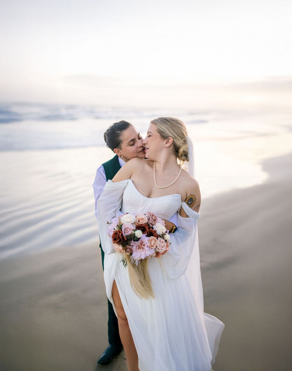 Romantic wedding couple embraces on a misty beach at sunset with pink and burgundy floral bouquet.