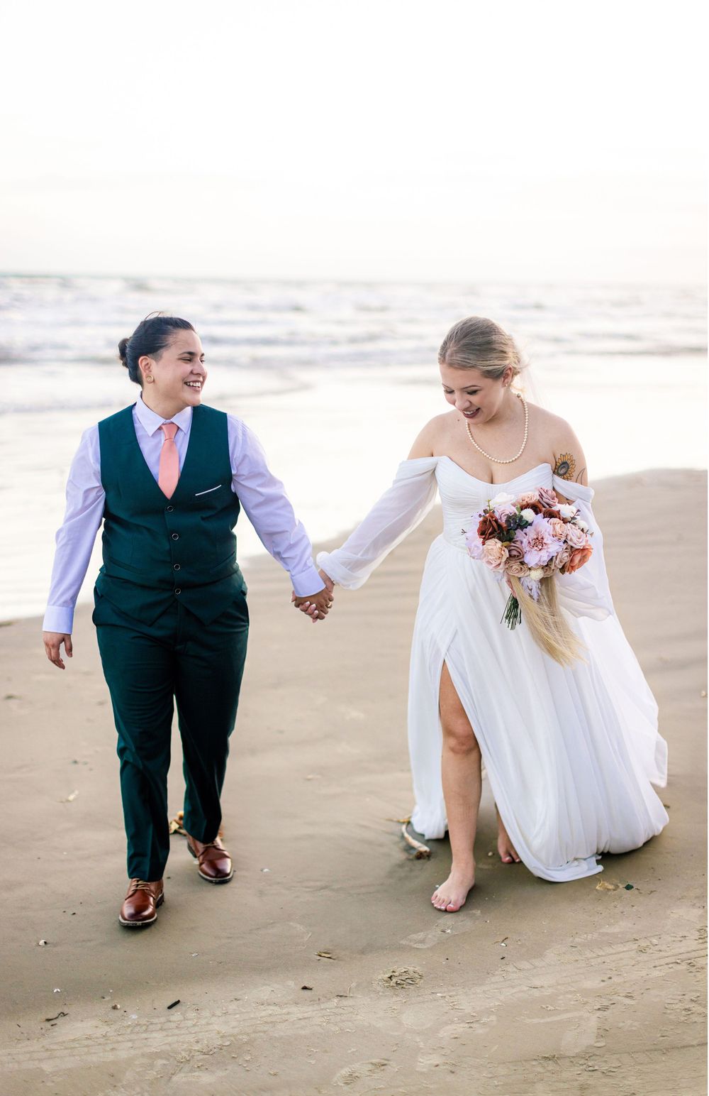 Newlyweds walk hand in hand on the beach at sunset with the bride holding a pink and white bouquet.
