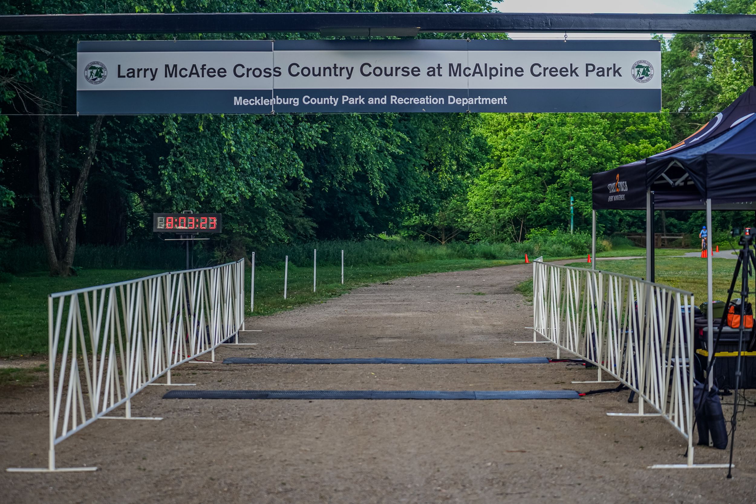 A dirt path with white metal barriers leads into a wooded area at Larry McAfee Cross Country Course.