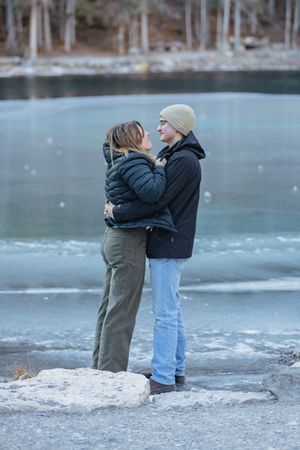 A couple in front of an ice covered lake embrace and stare lovingly at each other.