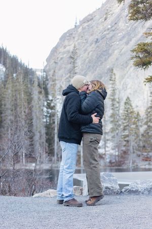 A couple kiss in front of a lake and mountain background. They embrace and the woman is on her tiptoes.
