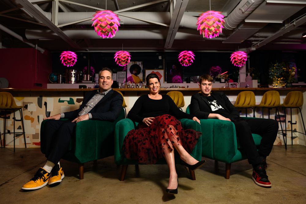 Three people seated on green velvet chairs under pink hanging lanterns in an industrial space.