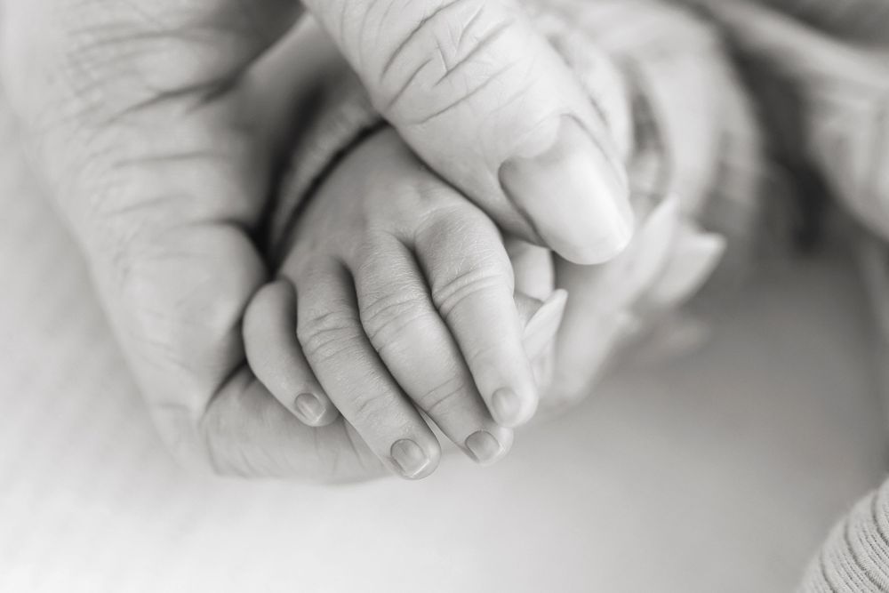 Black and white photo of a newborn baby's tiny hand gently held by an adult's larger hands.