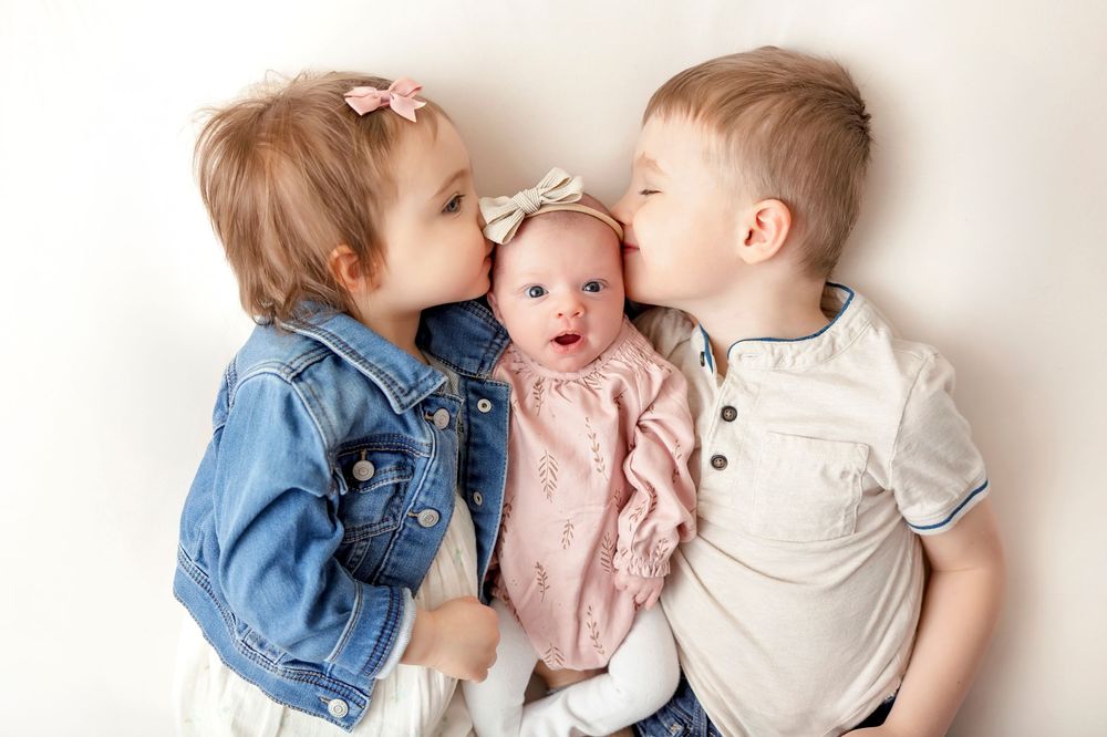 Siblings in casual clothes share sweet kisses with newborn baby.