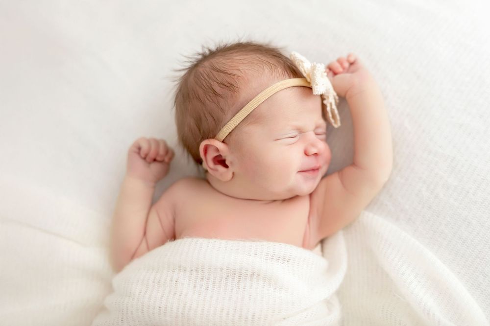 Sleeping newborn baby wearing a delicate headband lies peacefully on soft white blanket.