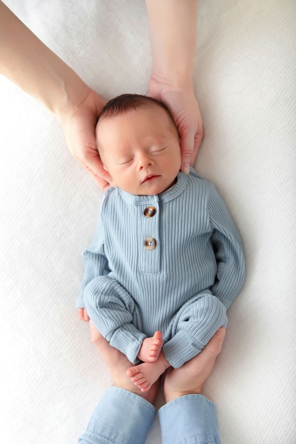 Sleeping newborn baby in blue ribbed romper cradled gently by parent hands on white background.