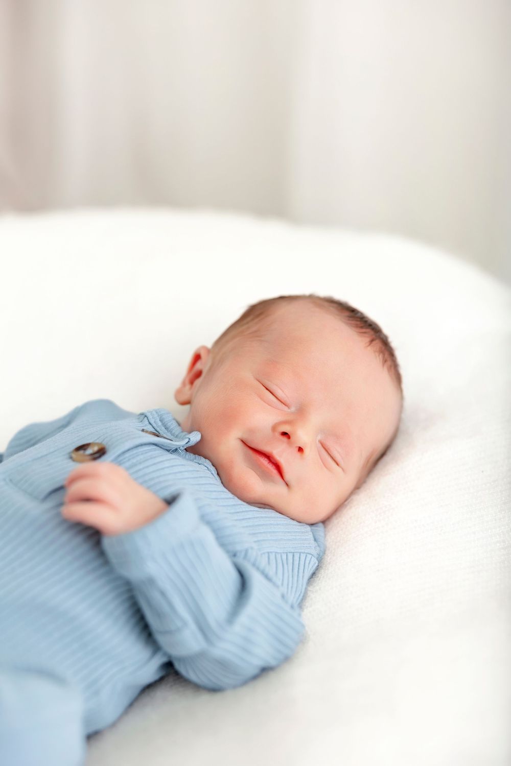 Newborn baby sleeping peacefully in a white basket, wearing a light blue ribbed onesie, with a gentle smile on face.