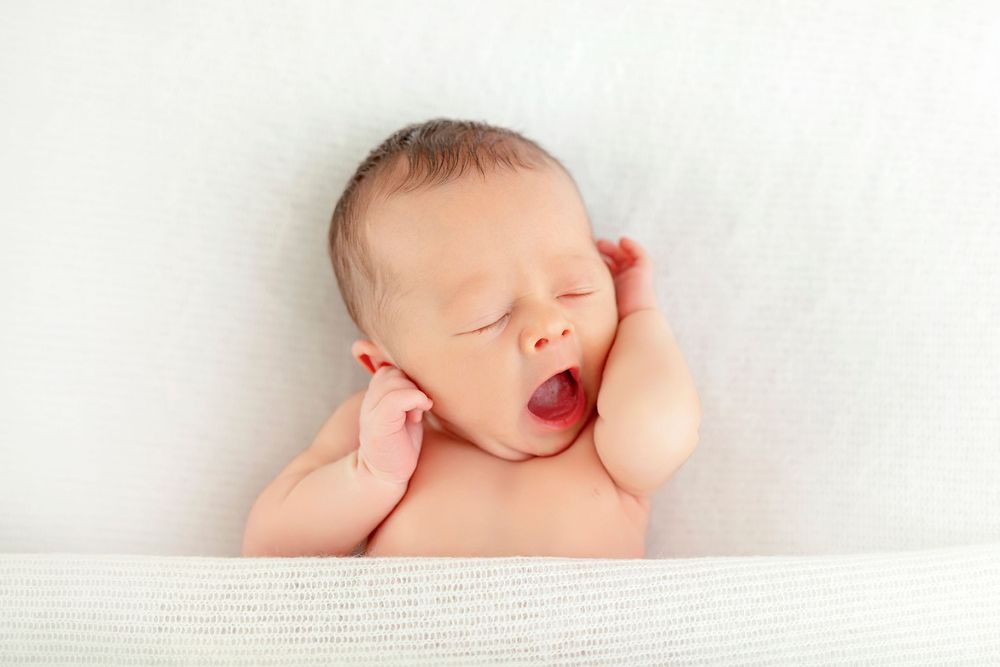 Newborn baby yawning with hands on cheeks, sleeping peacefully on a soft white blanket.