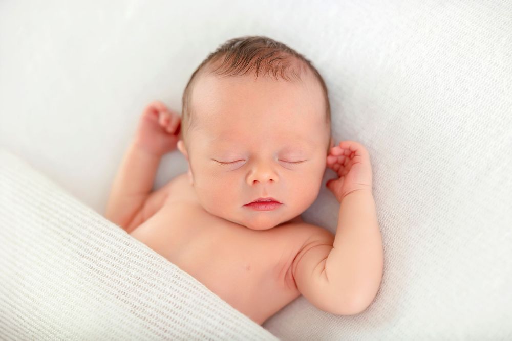 Peaceful newborn baby sleeping on a soft white blanket with tiny hands raised near face.