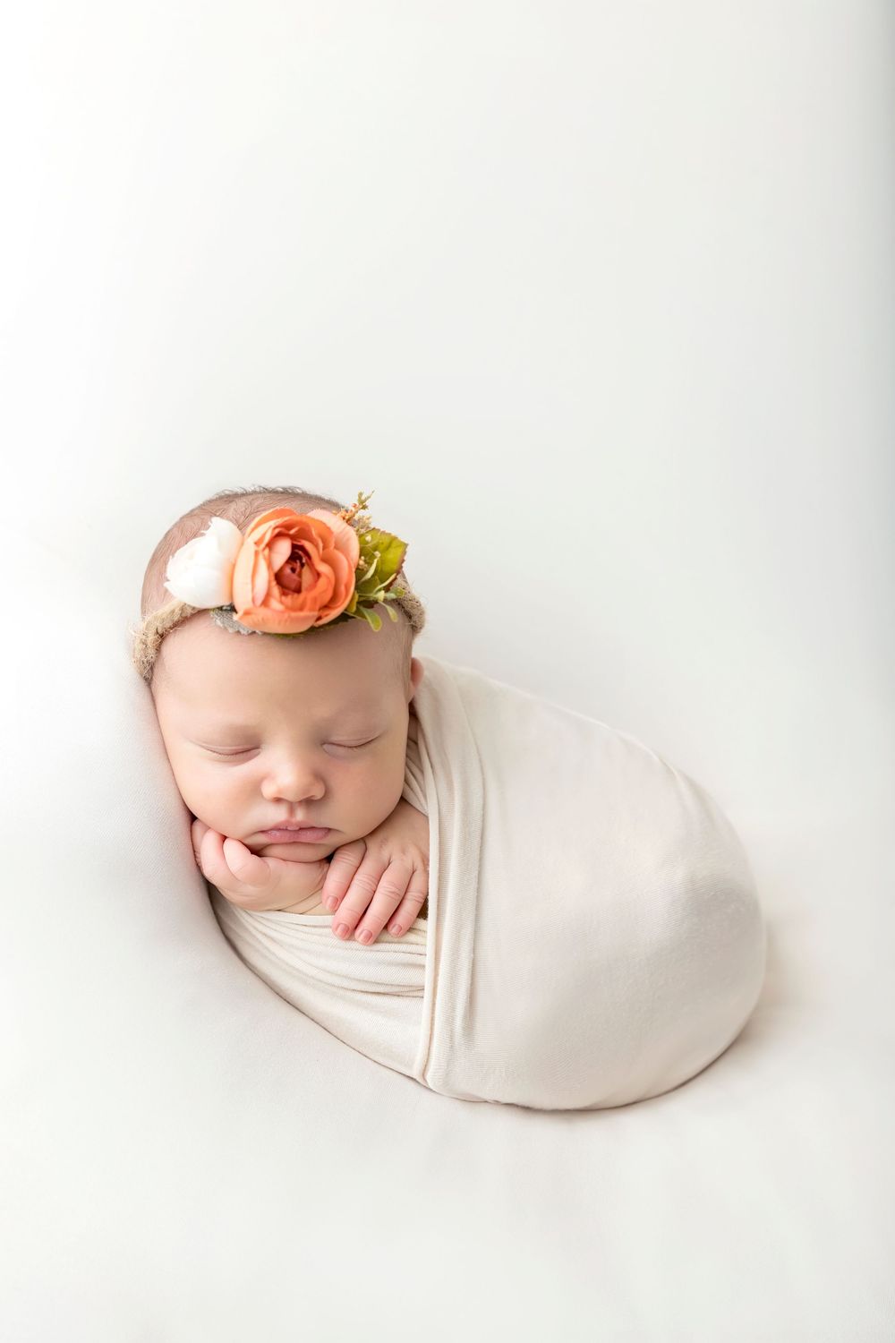 Sleeping newborn wrapped in cream fabric with delicate peach flower headband on white background.