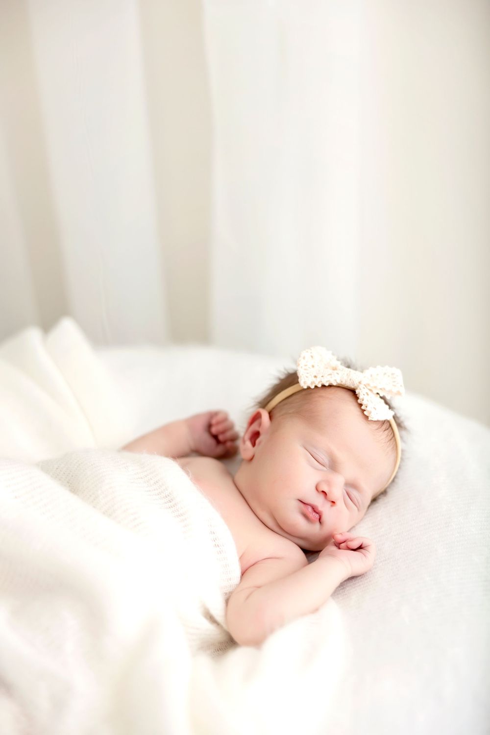 Sleeping newborn baby wearing white headband rests peacefully on soft white bedding in natural light.