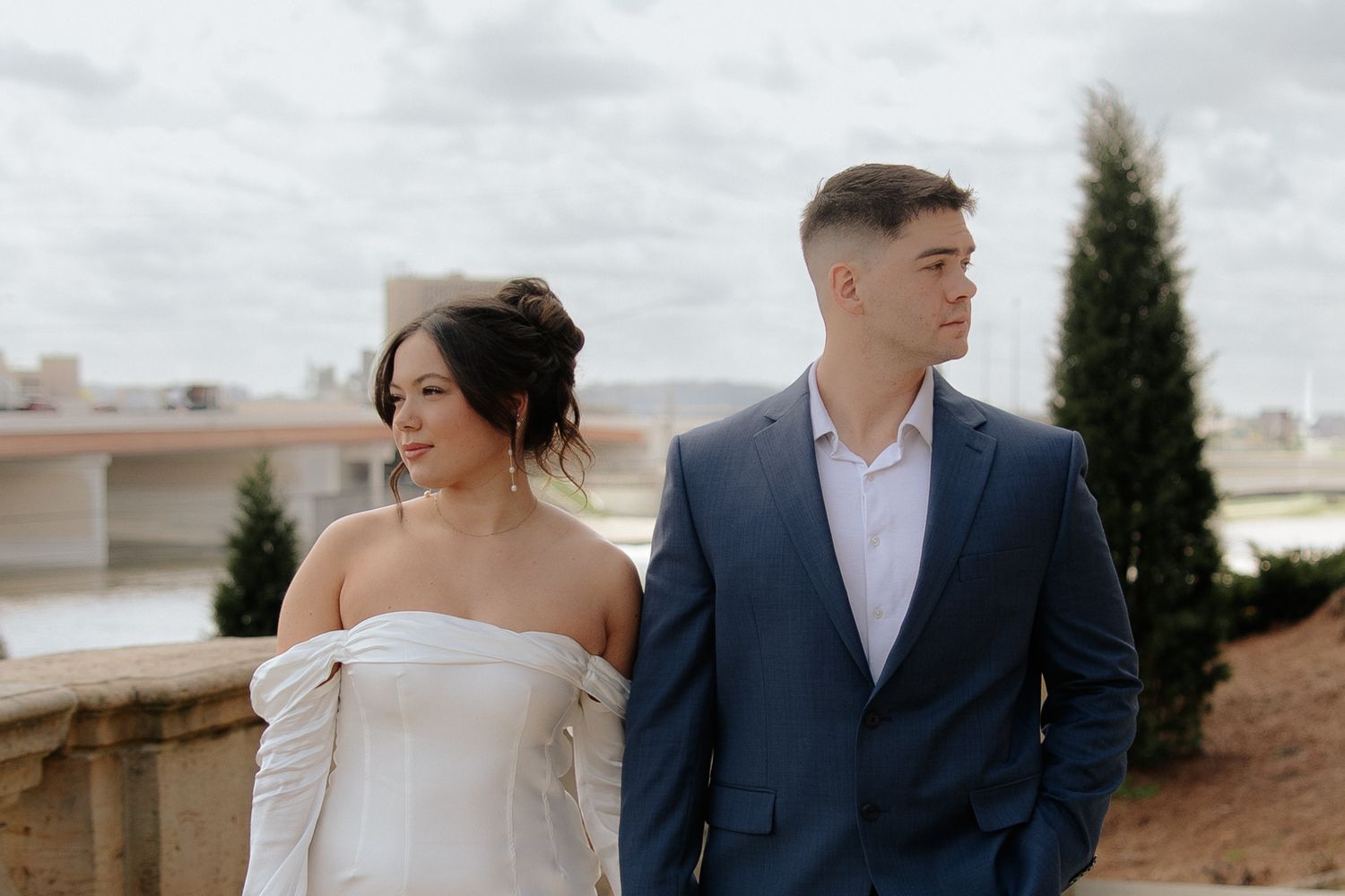 A couple stands together outdoors in formal attire, with the person in blue suit and another in off-shoulder white dress.