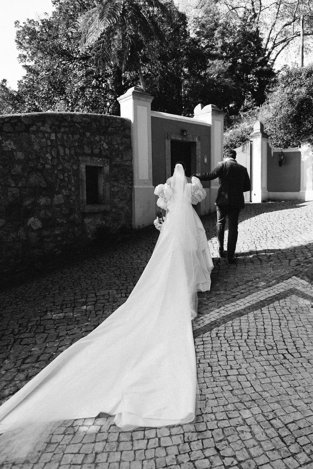A wedding couple walks along a cobblestone path with the bride's long white veil trailing behind them.