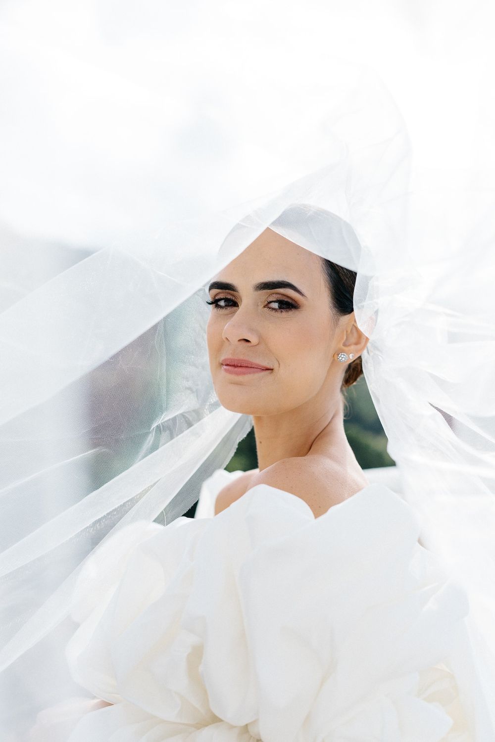A bride wearing a flowing white veil smiles softly in an ethereal portrait.