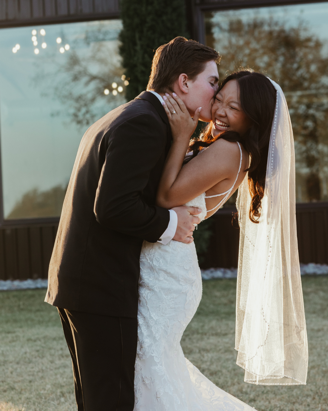 A wedding couple embraces during golden hour at sunset with a flowing veil catching the light.