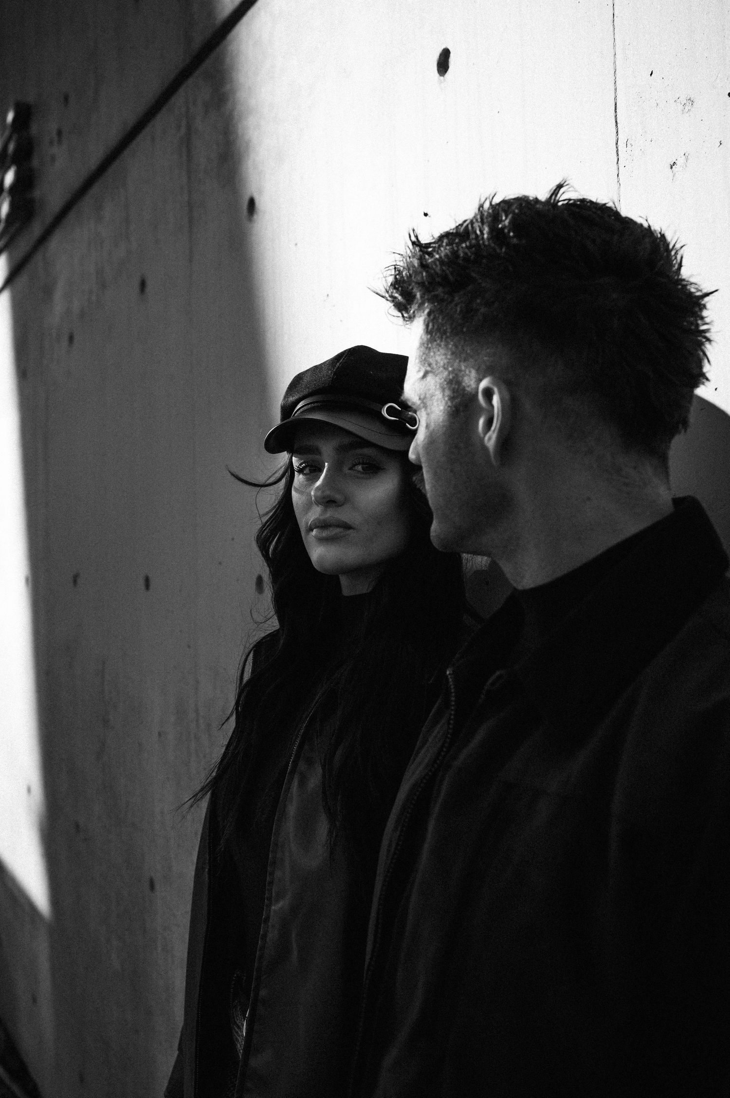 Black and white photo of a woman in a cap and a man in a jacket standing near a concrete wall.