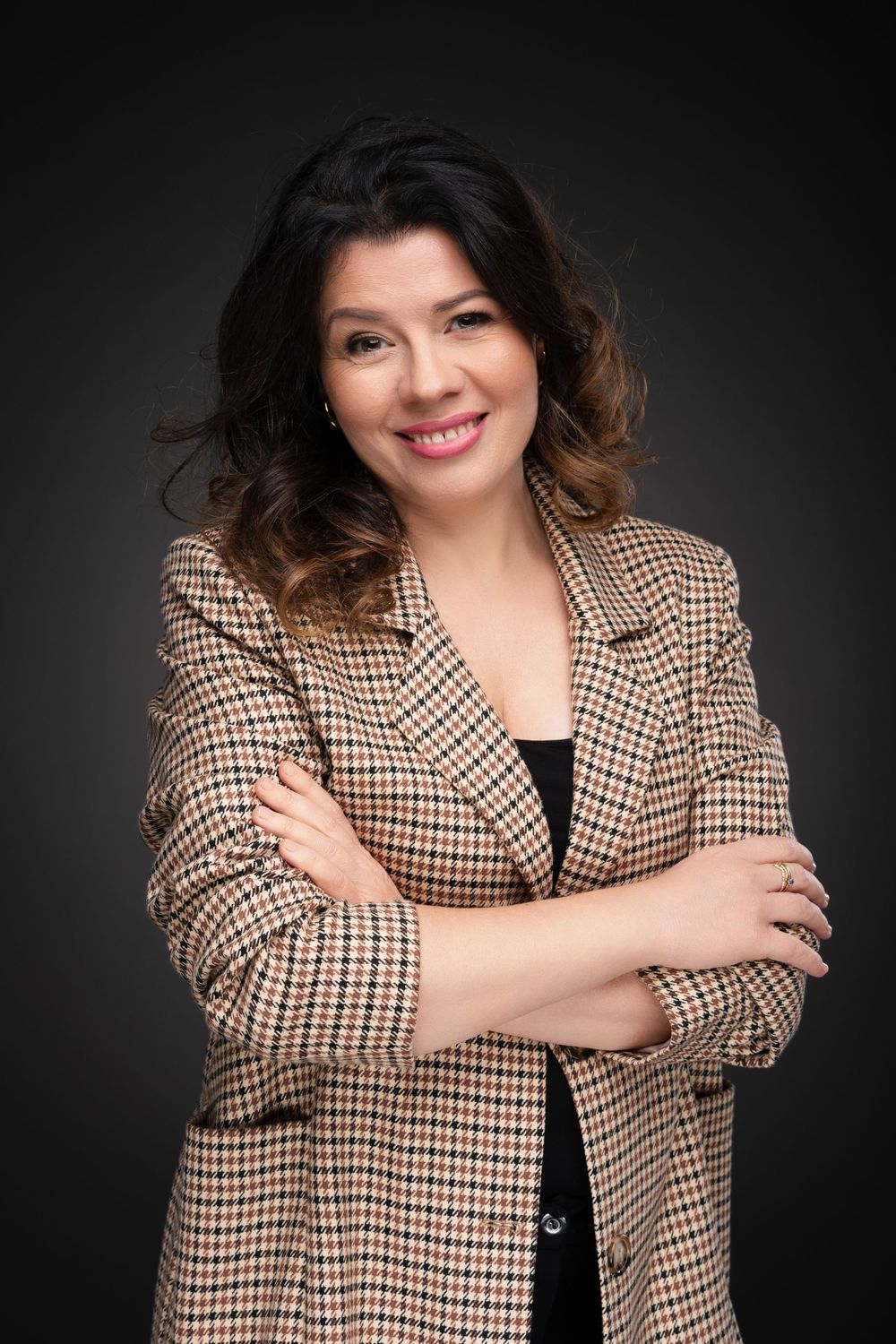 Professional studio portrait of person in plaid blazer with arms crossed against dark background.