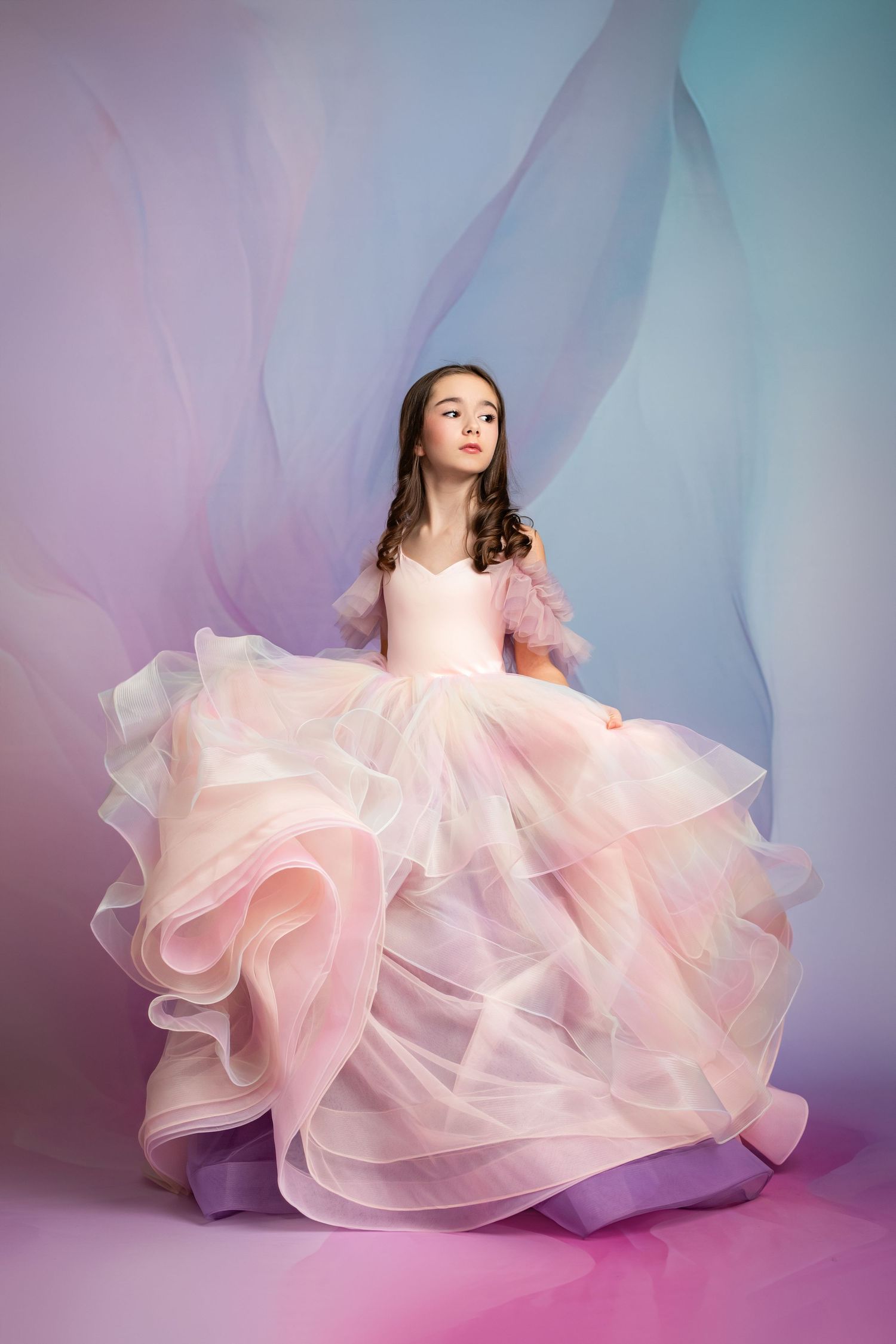 A young girl in a flowing pink princess gown with ruffled tulle layers poses against a dreamy purple and blue backdrop.