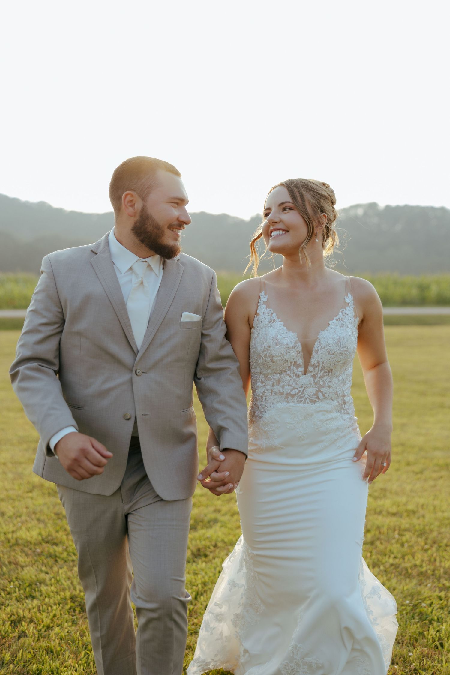 Newlyweds in grey suit and white dress running through sunny field.