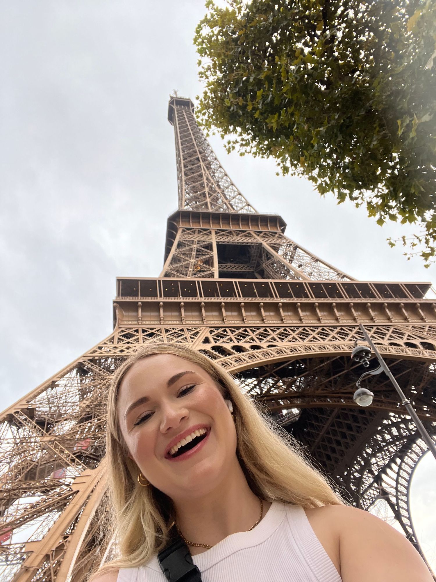 Smiling blonde woman taking a selfie in front of the Eiffel Tower in Paris on a cloudy day.