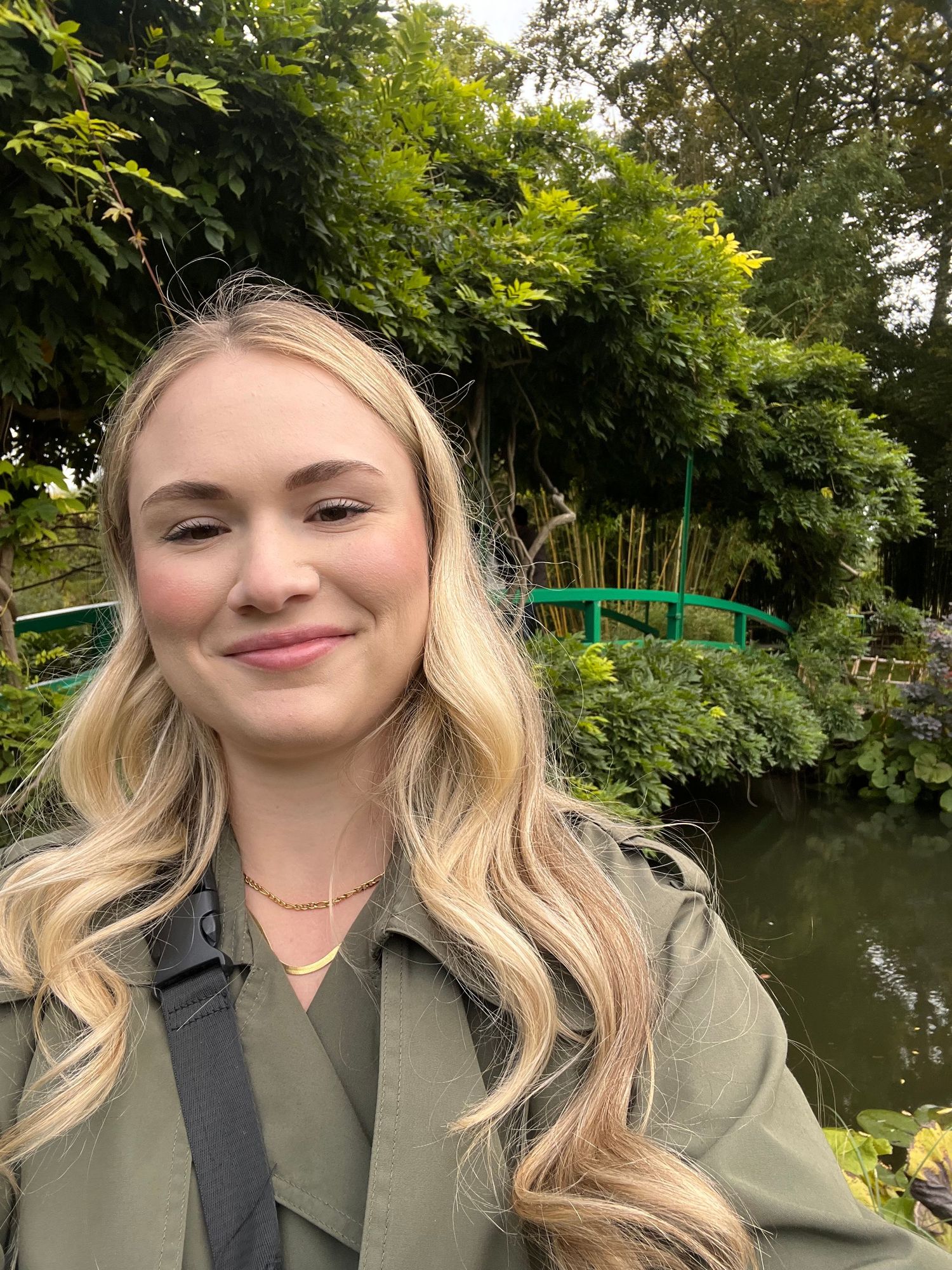Young blonde woman smiling outdoors near a green bridge and lush garden waterway on a cloudy day.